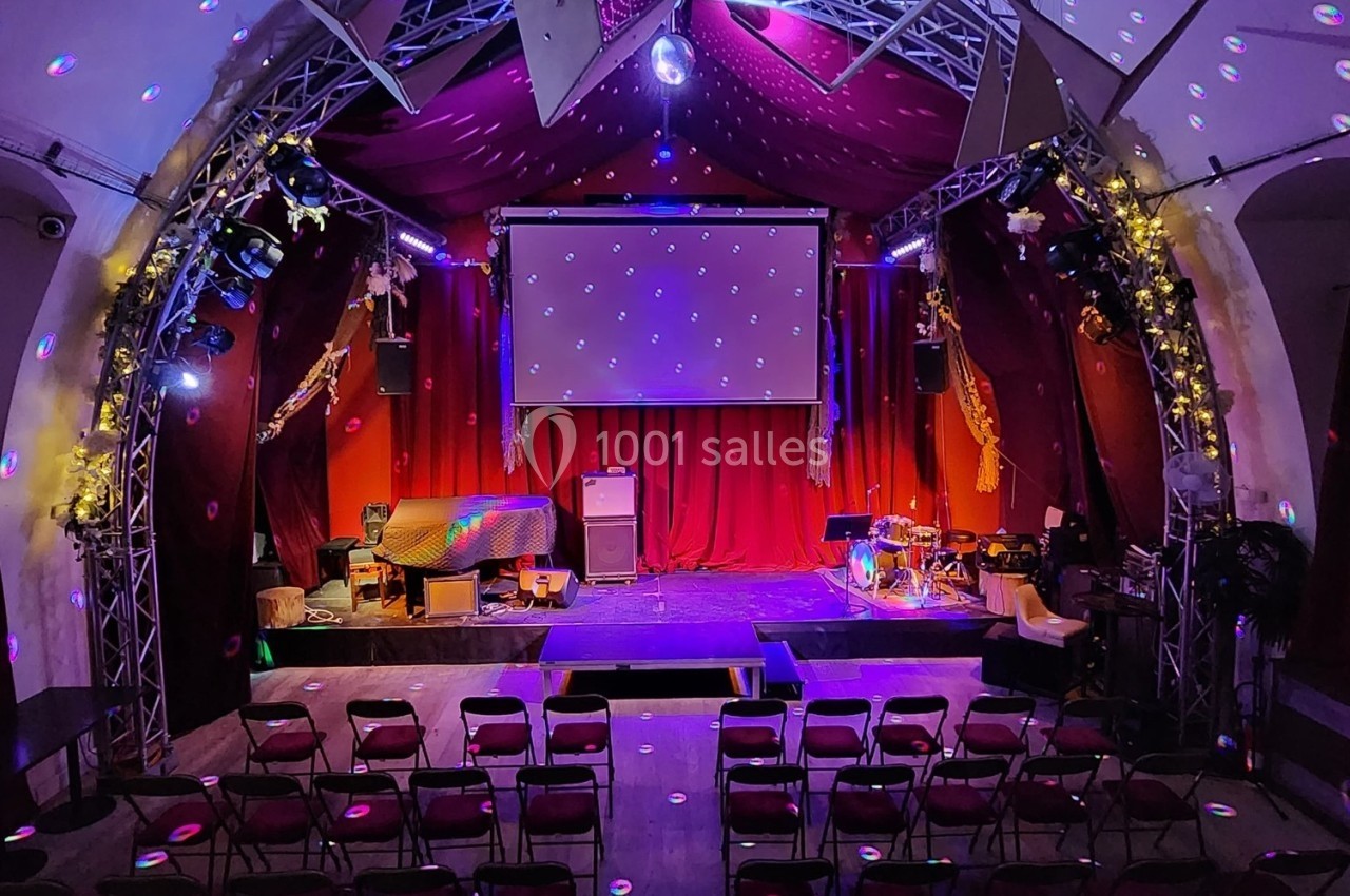 Salle de spectacle avec scène éclairée, rideaux rouges, projecteurs colorés et chaises disposées en rangées.