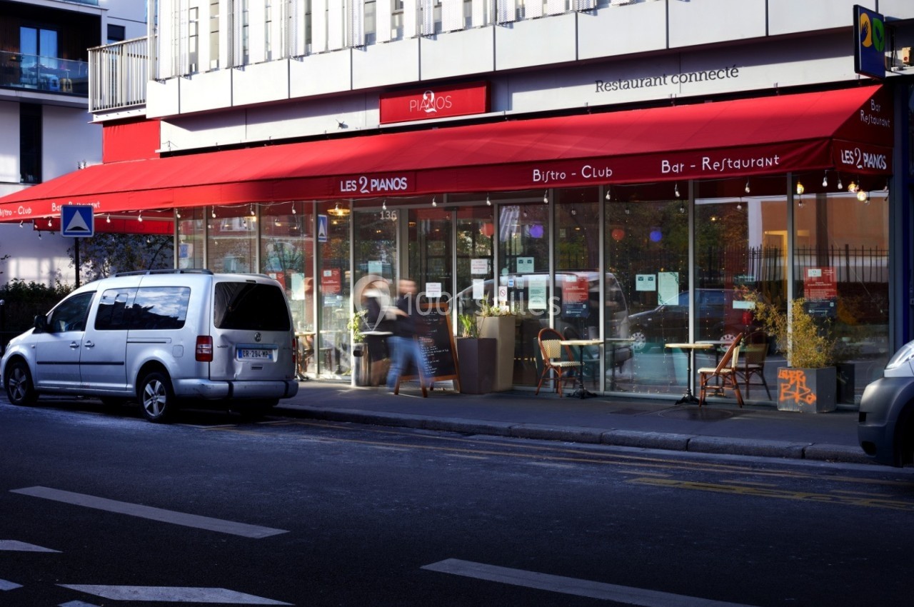Façade d'un restaurant avec terrasse, abritée par un large store rouge, situé en bordure de rue.