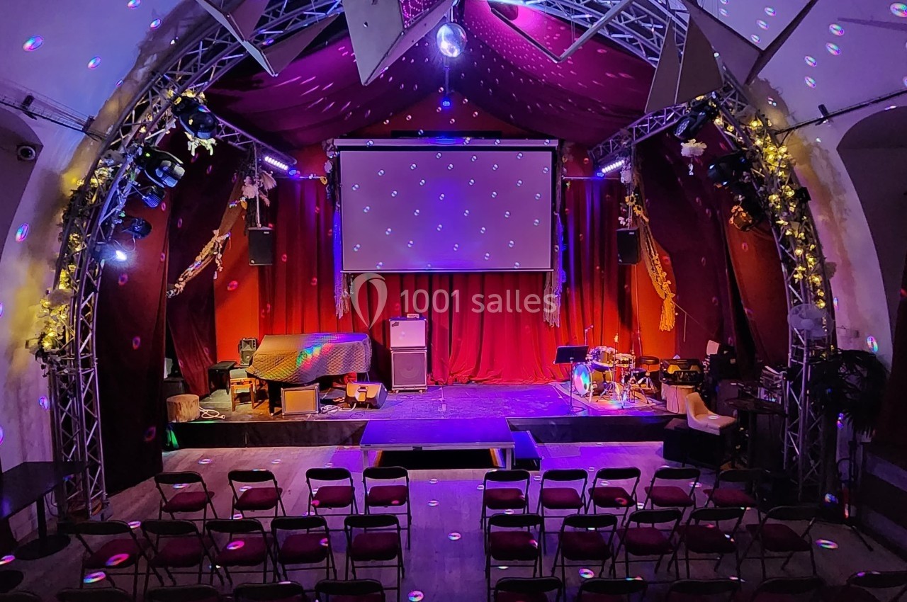 Salle de spectacle avec scène éclairée, rideaux rouges, projecteurs colorés et rangées de chaises vides.