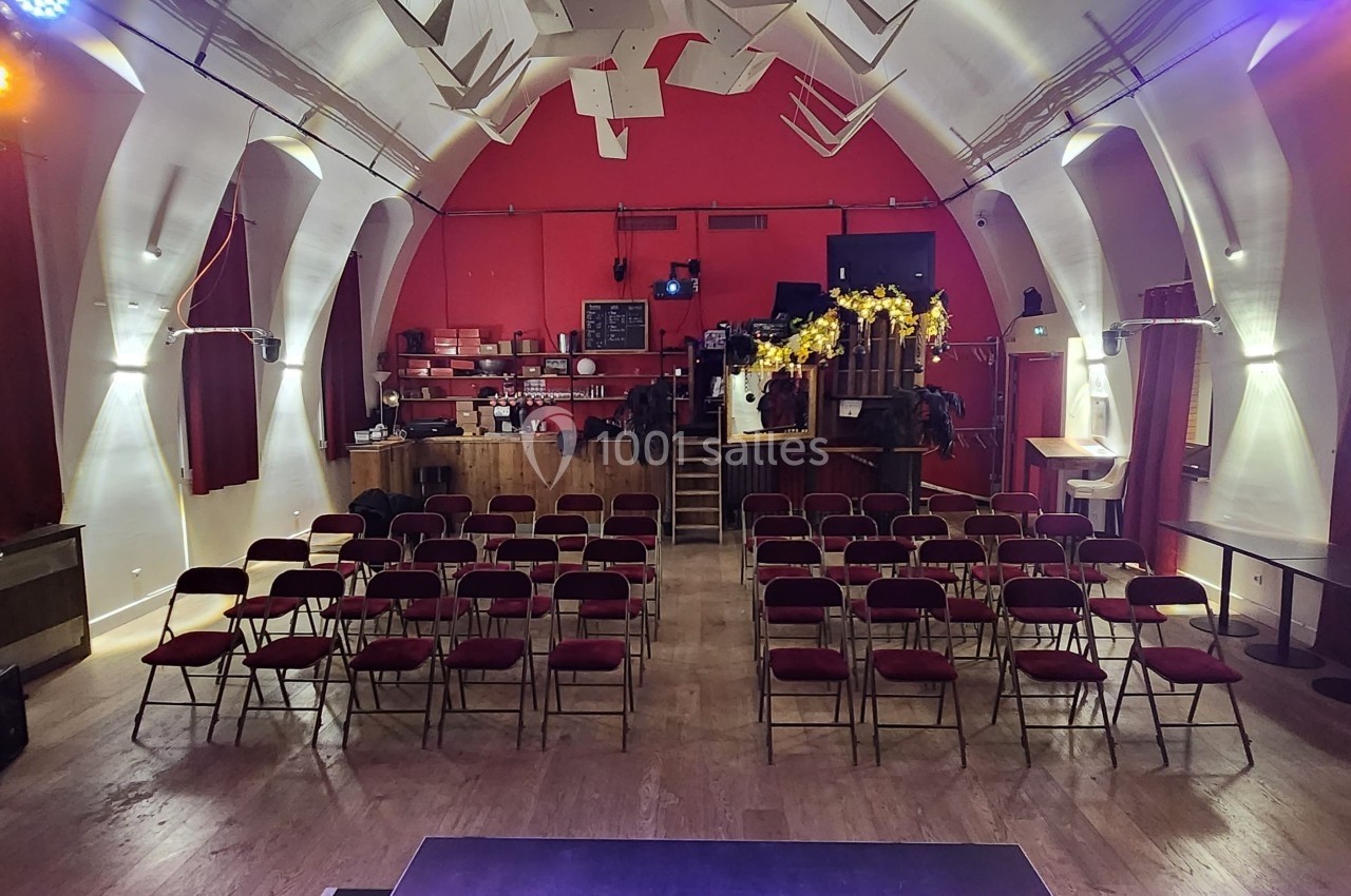 Salle avec des rangées de chaises rouges face à une scène, décorée de lumières et d'un bar en arrière-plan.