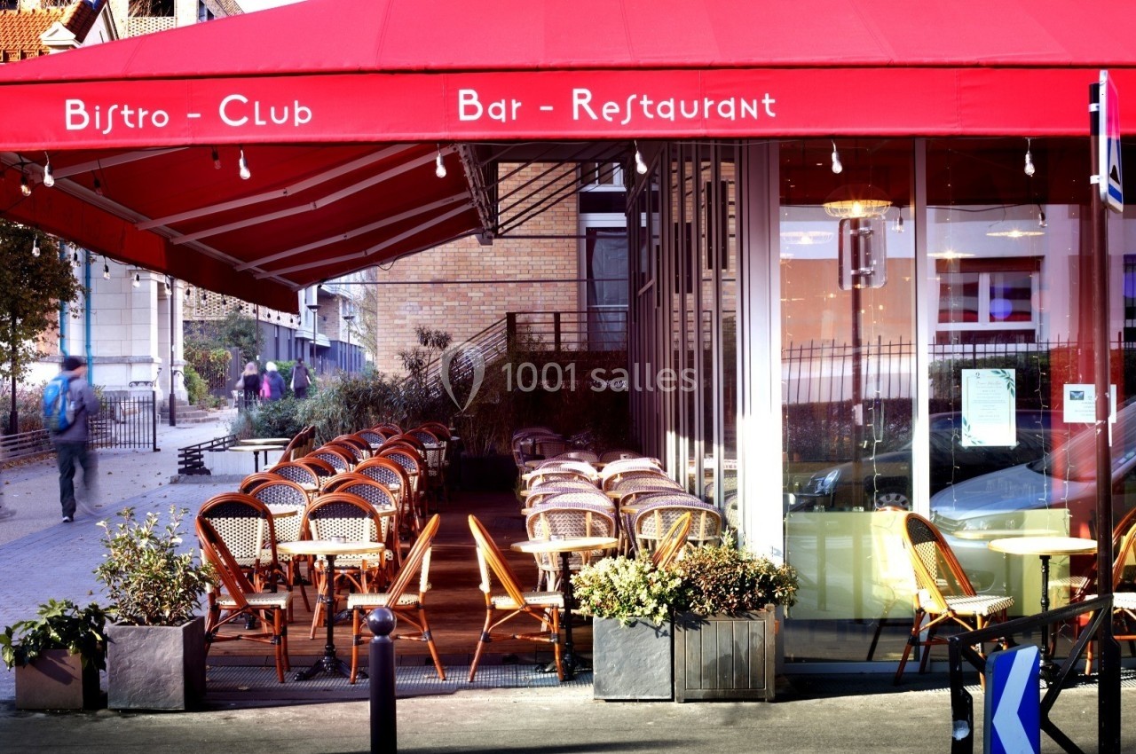 Terrasse extérieure d'un bistro avec des chaises en osier sous un auvent rouge, située près d'une rue piétonne.