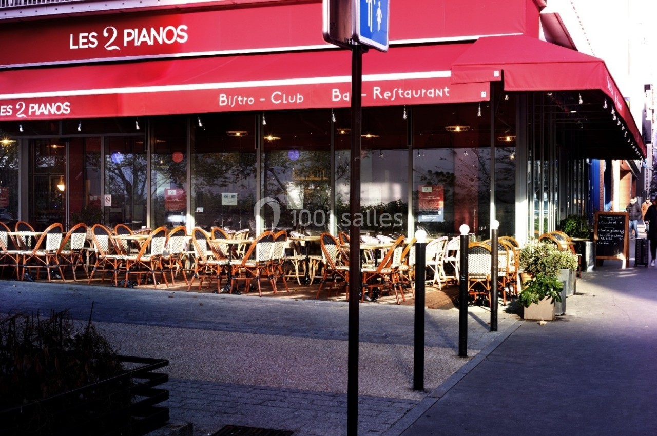 Terrasse d'un restaurant avec des tables et chaises en osier sous un auvent rouge, située en bord de trottoir.