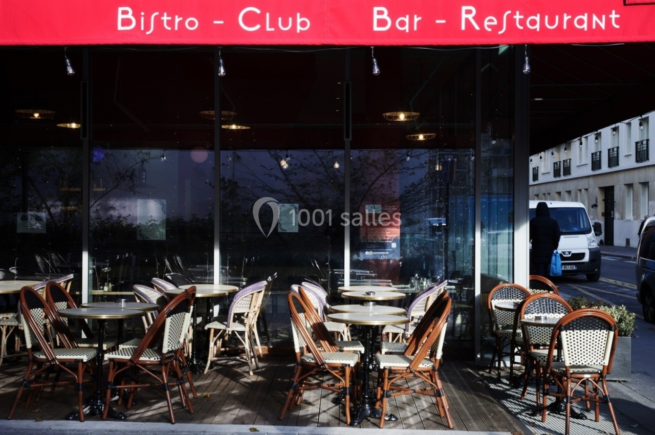 Terrasse extérieure d'un bistro avec des tables et chaises en osier sous un auvent rouge, rue en arrière-plan.