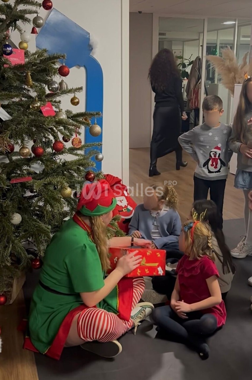 Une personne déguisée en lutin offre un cadeau à des enfants assis près d'un sapin de Noël décoré.