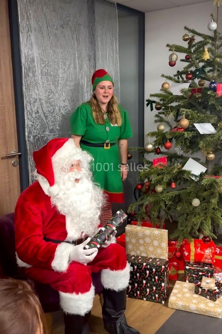 Un Père Noël assis tenant un cadeau, accompagné d'une assistante en tenue d'elfe près d'un sapin décoré.