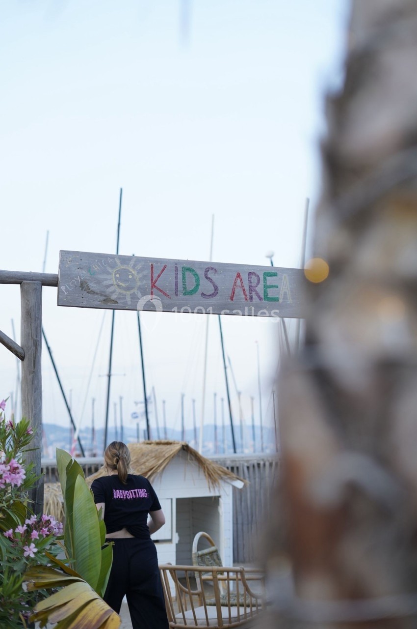 Panneau ’Kids Area’ en bois près d'une aire de jeux, avec une femme de dos portant un t-shirt ’Babysitting’.