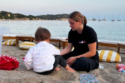 Panneau ’Kids Area’ en bois près d'une aire de jeux, avec une femme de dos portant un t-shirt ’Babysitting’.