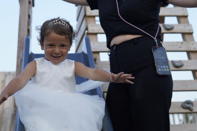 Panneau ’Kids Area’ en bois près d'une aire de jeux, avec une femme de dos portant un t-shirt ’Babysitting’.