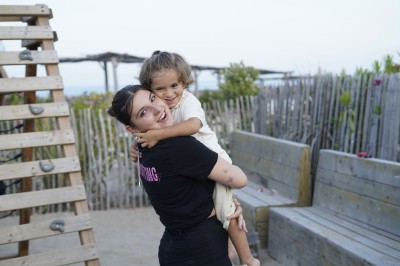 Panneau ’Kids Area’ en bois près d'une aire de jeux, avec une femme de dos portant un t-shirt ’Babysitting’.