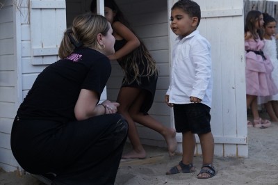 Panneau ’Kids Area’ en bois près d'une aire de jeux, avec une femme de dos portant un t-shirt ’Babysitting’.
