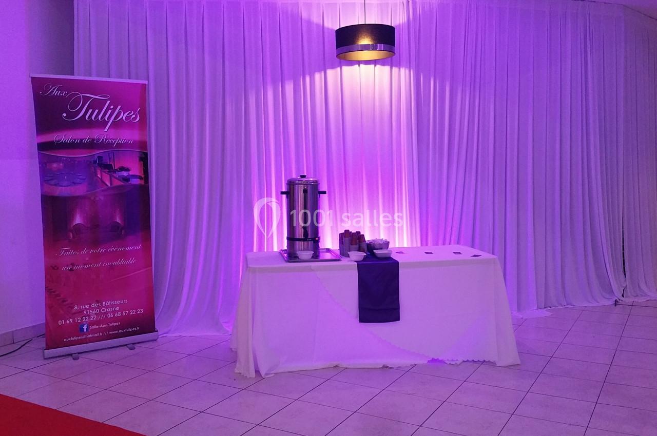Table dressée avec une nappe blanche, urne à café et accessoires, devant un rideau blanc éclairé de lumière violette.