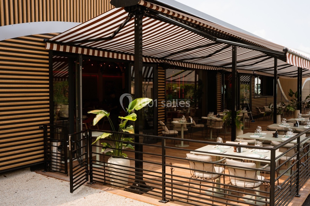 Terrasse d'un restaurant avec des tables dressées, des plantes en pot et des stores rayés noirs et blancs.