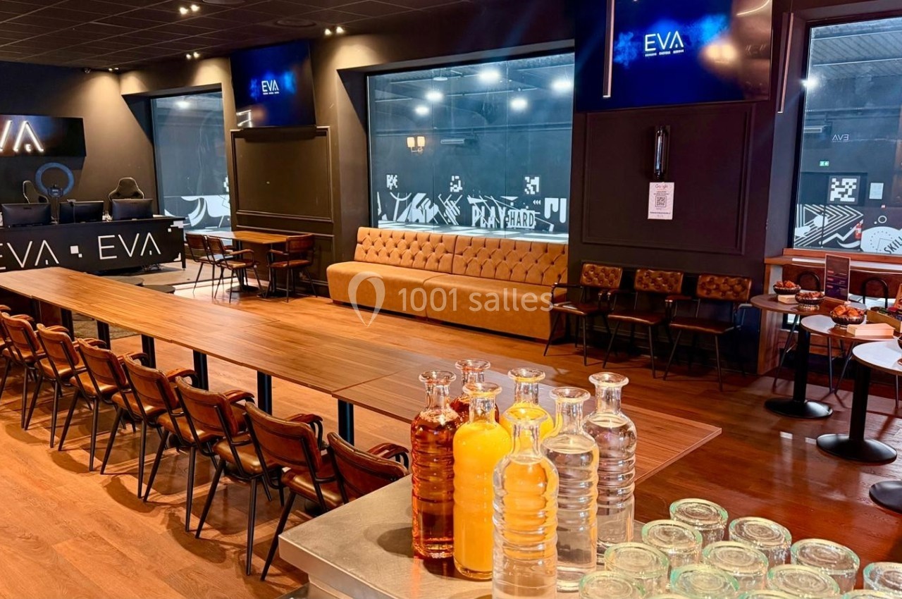 Salle spacieuse avec une longue table en bois, chaises, canapés, écrans et un comptoir avec boissons et carafes.