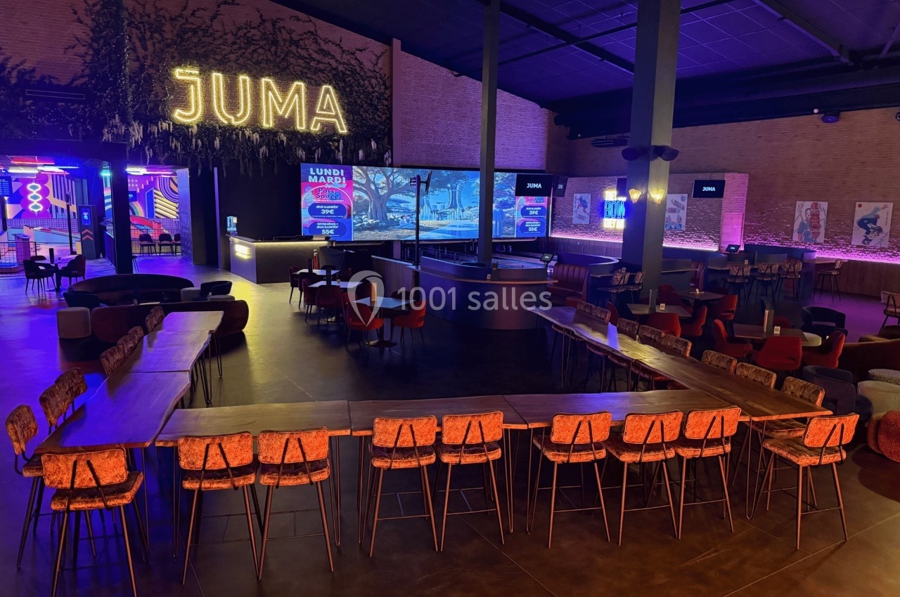 Salle moderne avec tables, chaises et bar central, éclairée par des néons et un grand écran en arrière-plan.