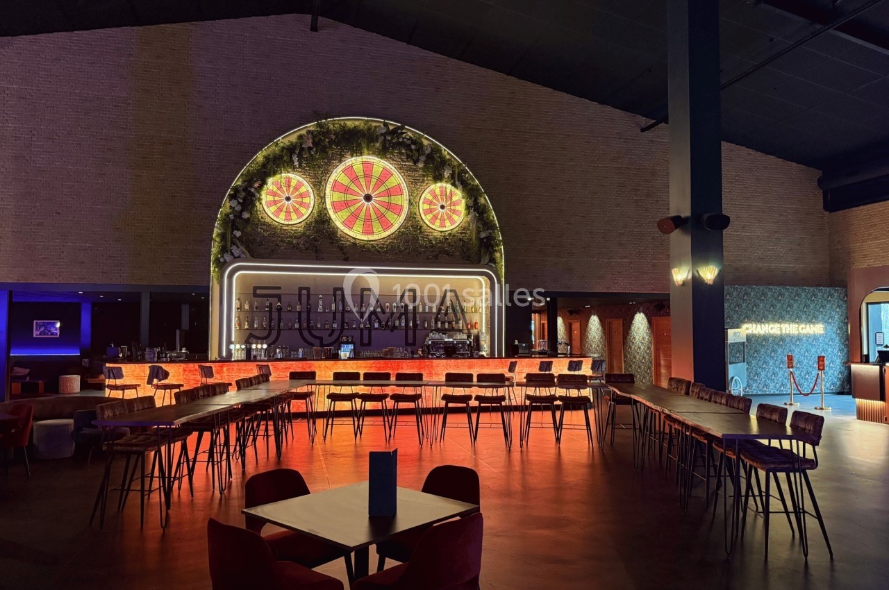 Intérieur d'un bar avec un long comptoir éclairé, des tables et des chaises, et une décoration lumineuse au mur.