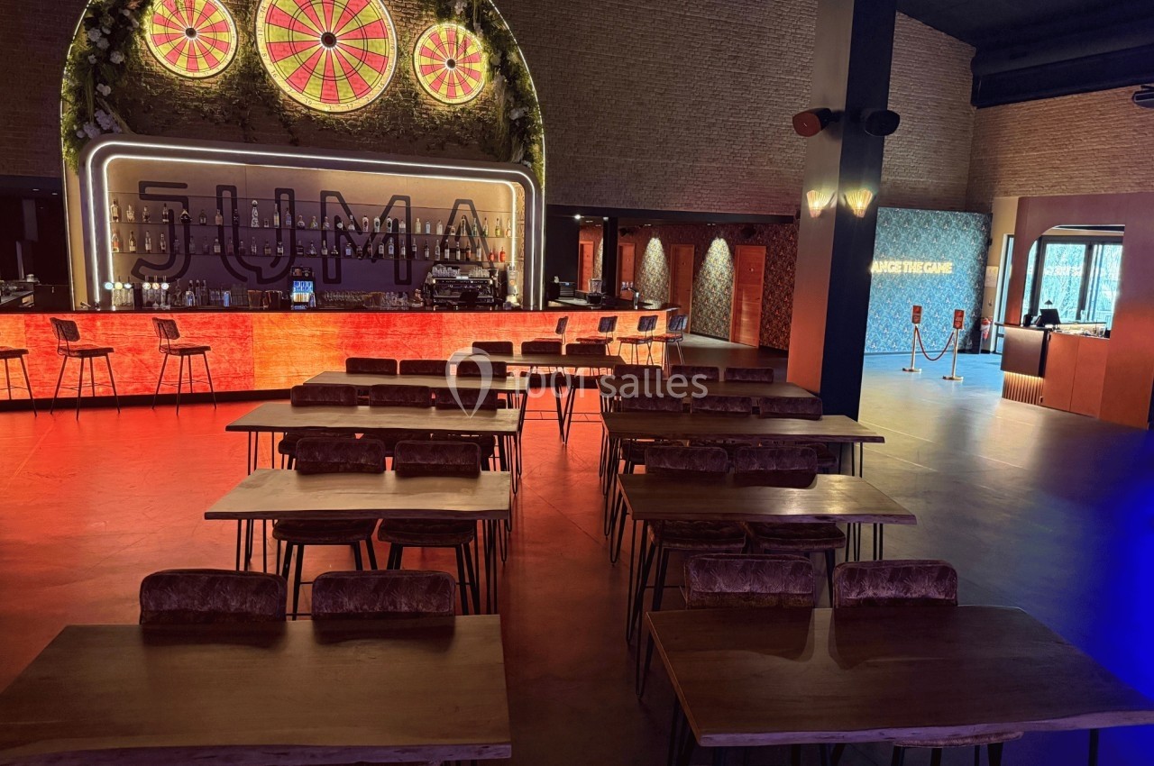 Salle avec tables en bois, chaises rembourrées, bar éclairé en arrière-plan et décorations lumineuses.