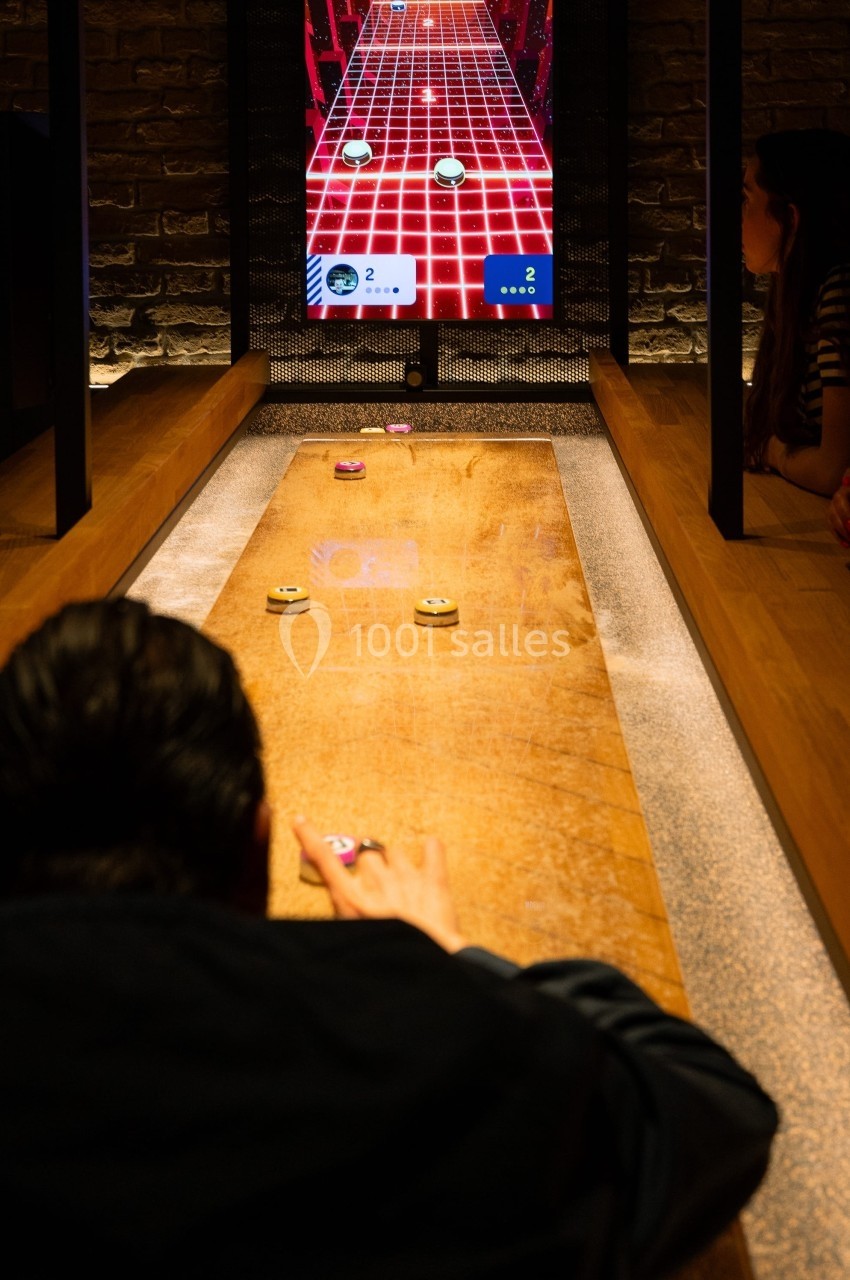 Personnes jouant à un jeu interactif de palets sur une table en bois, avec un écran affichant le score en arrière-plan.