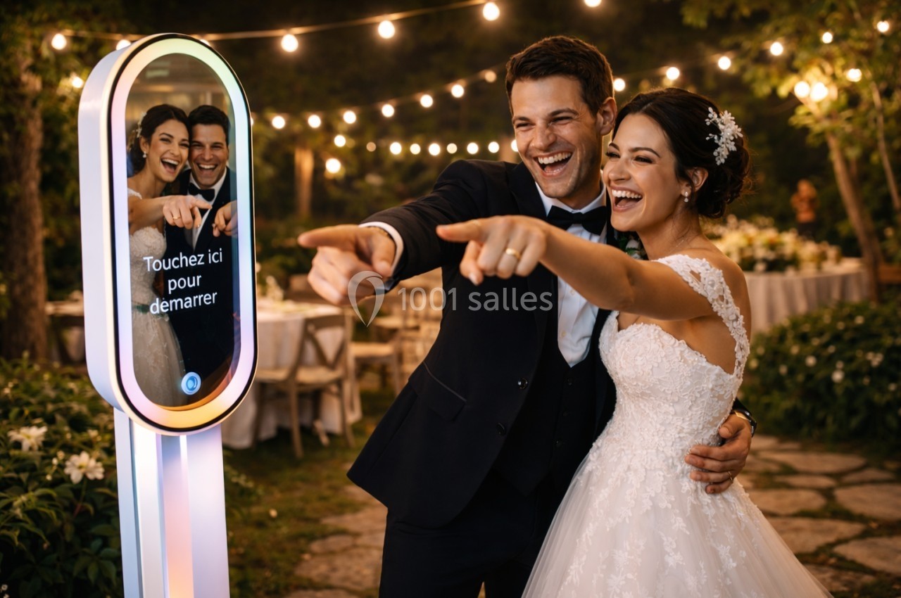 Un couple souriant en tenue de mariage pose devant une borne photo interactive lors d'une réception en extérieur.