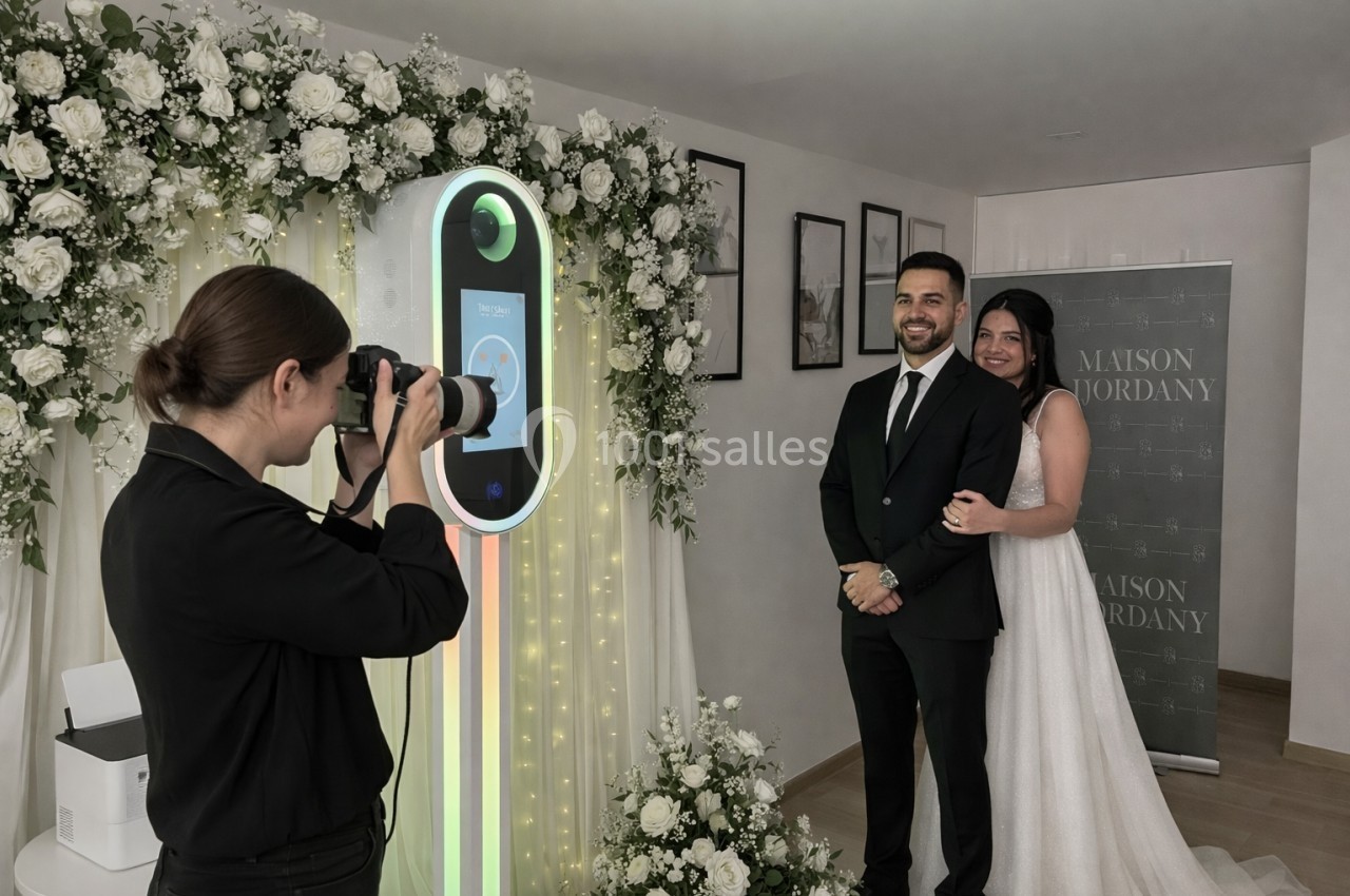 Un couple en tenue de mariage pose devant un photobooth décoré de fleurs, photographié par une femme.