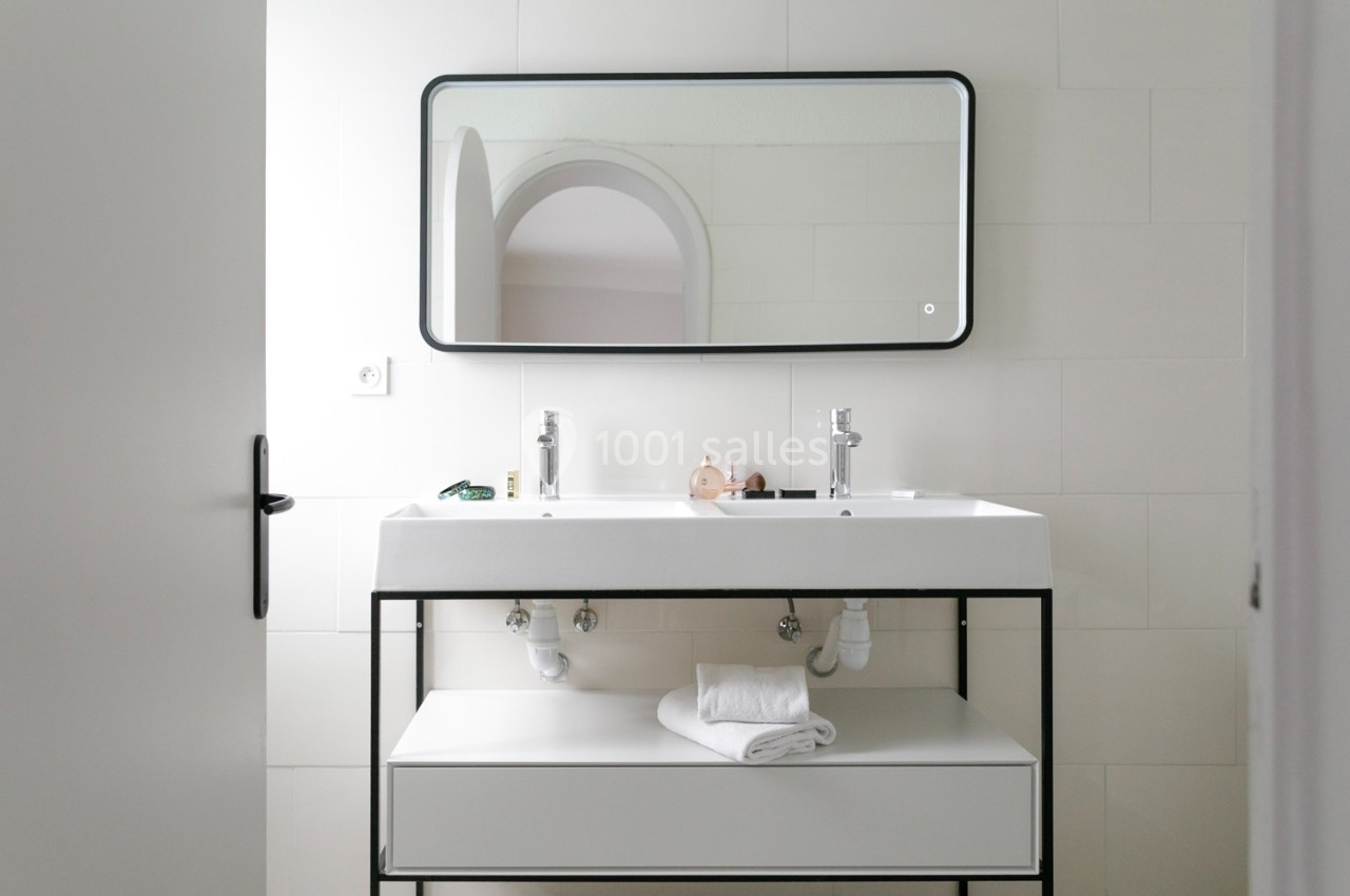 Lavabo double avec miroir rectangulaire, serviette pliée sur une étagère et mur carrelé blanc en arrière-plan.