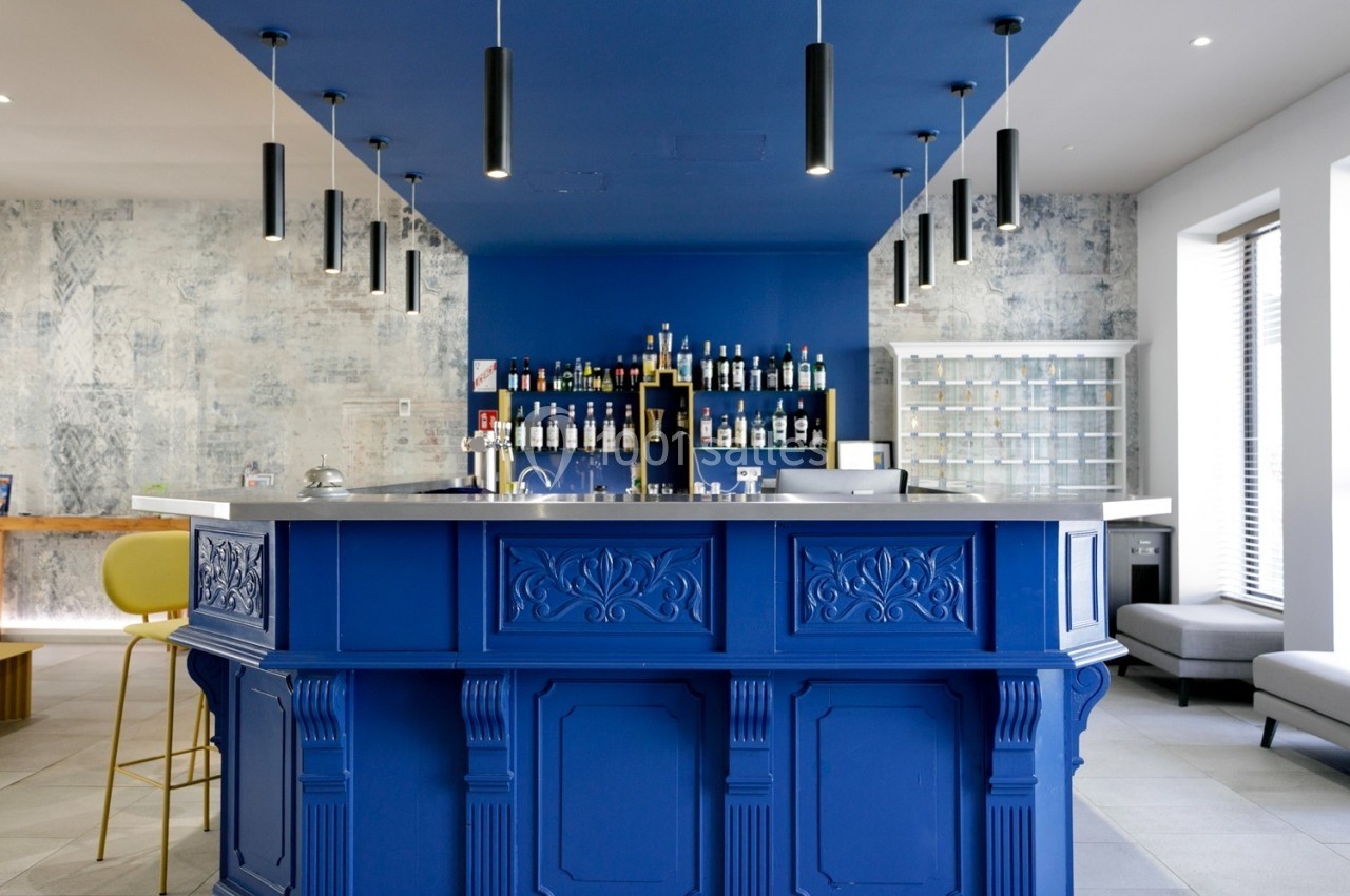 Comptoir de bar bleu avec étagères de bouteilles en arrière-plan, éclairage suspendu et décor moderne.