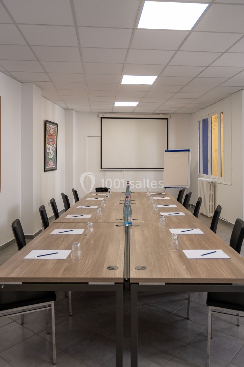 Salle de réunion avec une grande table rectangulaire, des chaises, un tableau blanc et un écran de projection.