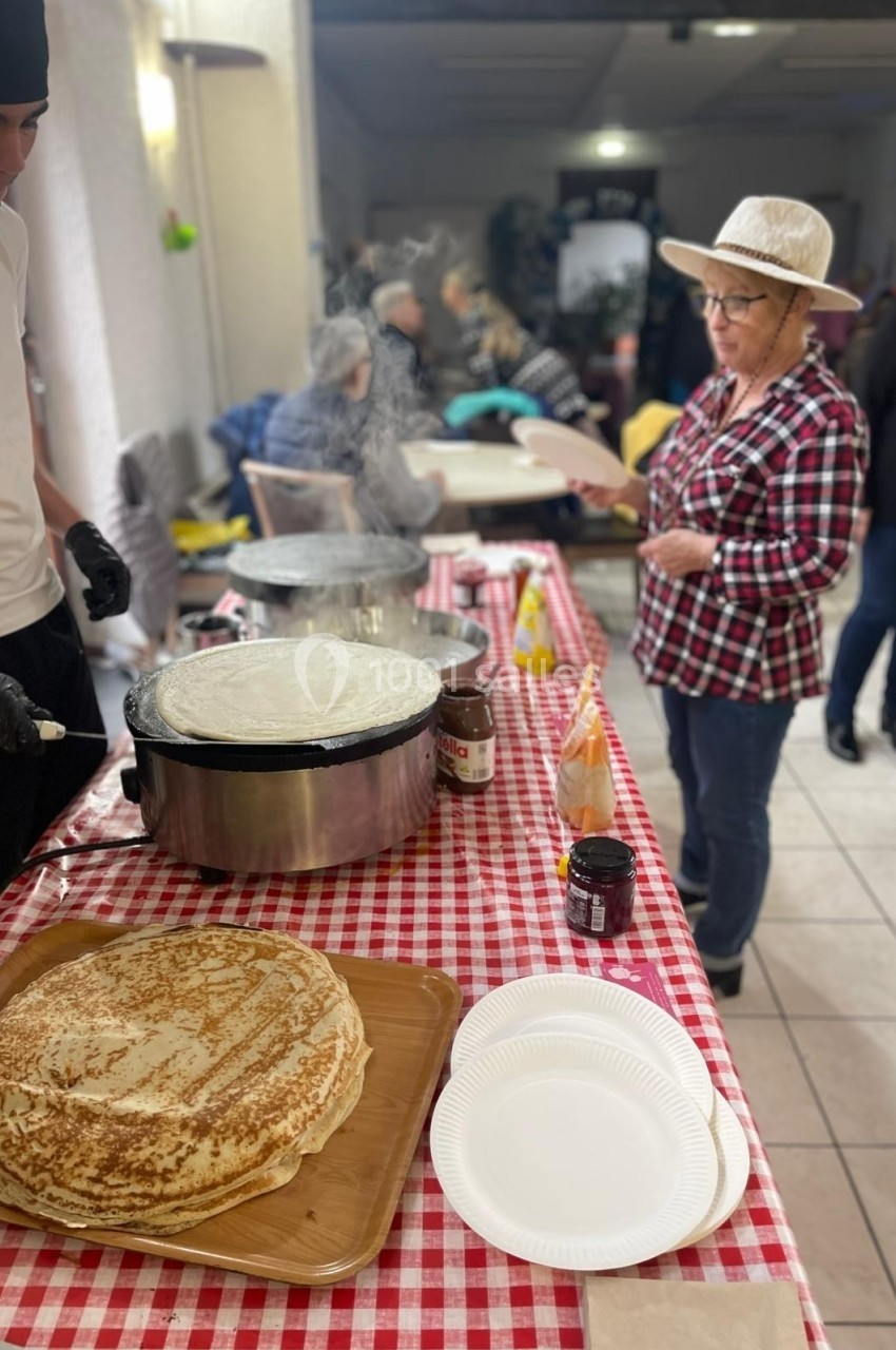 Un stand de crêpes avec un cuisinier et une femme en chapeau, dans une salle animée.