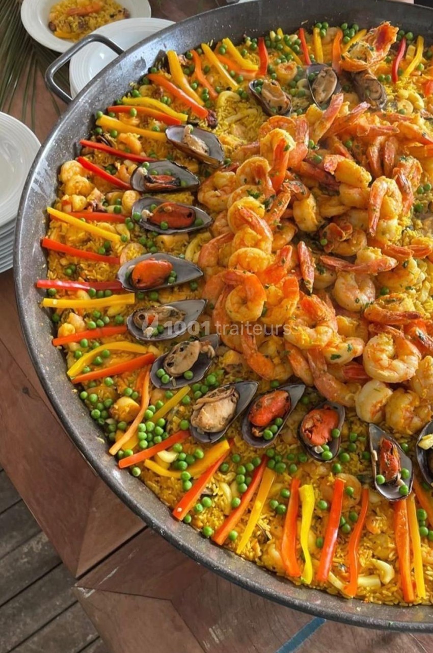 Grande poêle de paella garnie de crevettes, moules, légumes colorés et riz safrané, présentée sur une table en bois.