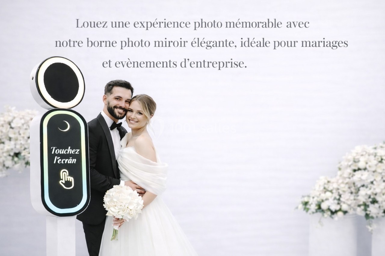 Un couple de mariés souriant pose près d'une borne photo miroir dans un décor lumineux et élégant.
