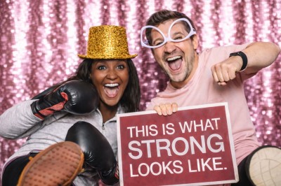 Un couple souriant en tenue de mariage pose devant une borne photo interactive lors d'une réception en extérieur.