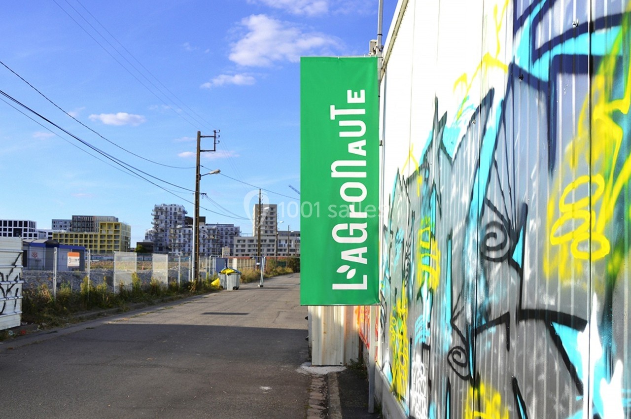 Banderole verte ’L'Agronaute’ accrochée à une clôture graffée, avec des bâtiments en arrière-plan.