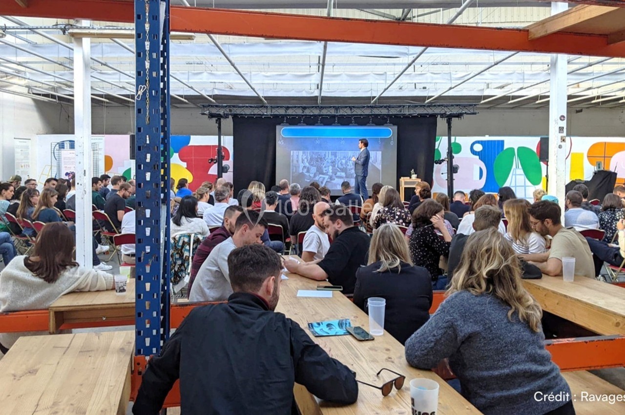 Un conférencier s'adresse à un public assis dans un espace lumineux avec des tables en bois et des décorations colorées.