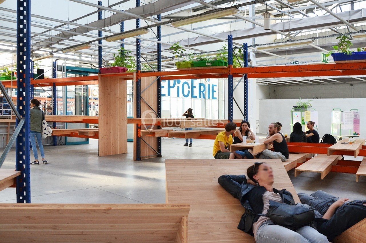 Des personnes discutent et se détendent sur des bancs en bois dans un espace intérieur lumineux avec une épicerie en arrière…