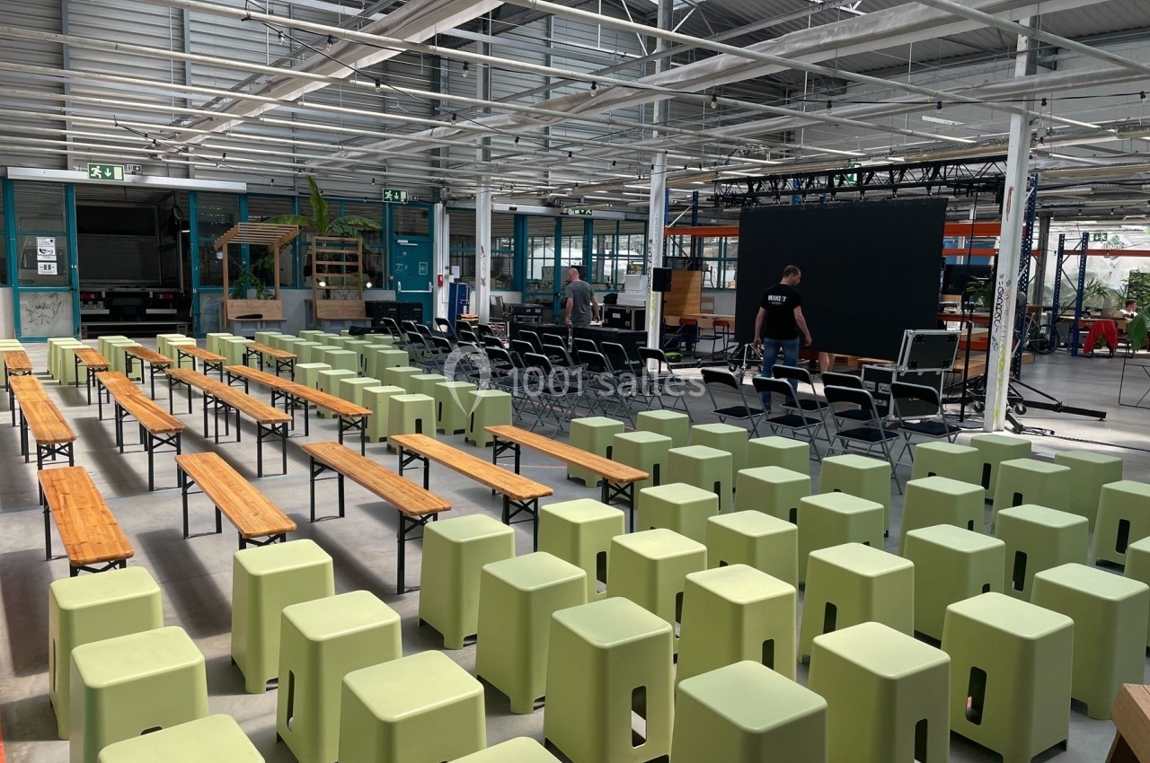Salle lumineuse aménagée avec des bancs en bois, des tabourets verts et un écran noir pour un événement.