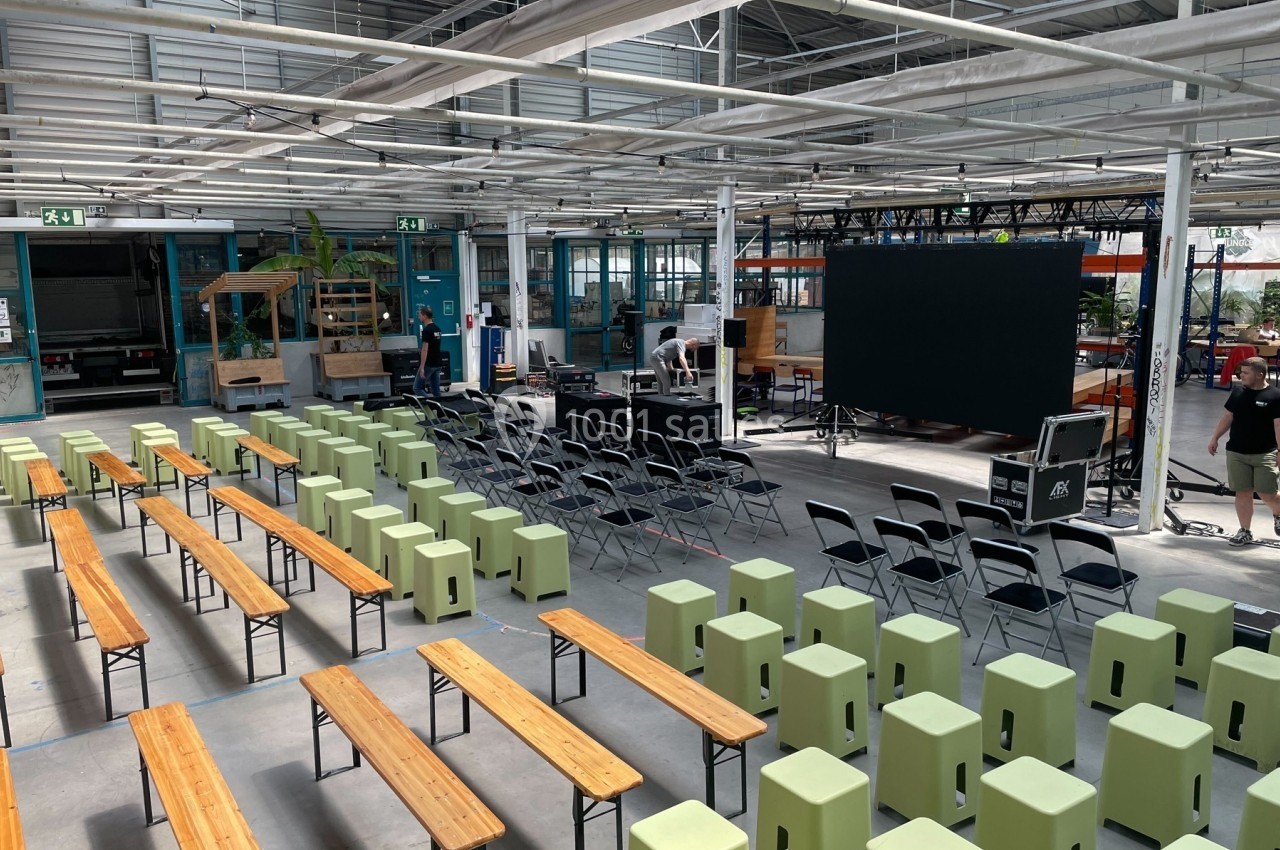 Salle industrielle aménagée avec des bancs, des chaises et un écran géant, prête pour un événement ou une présentation.
