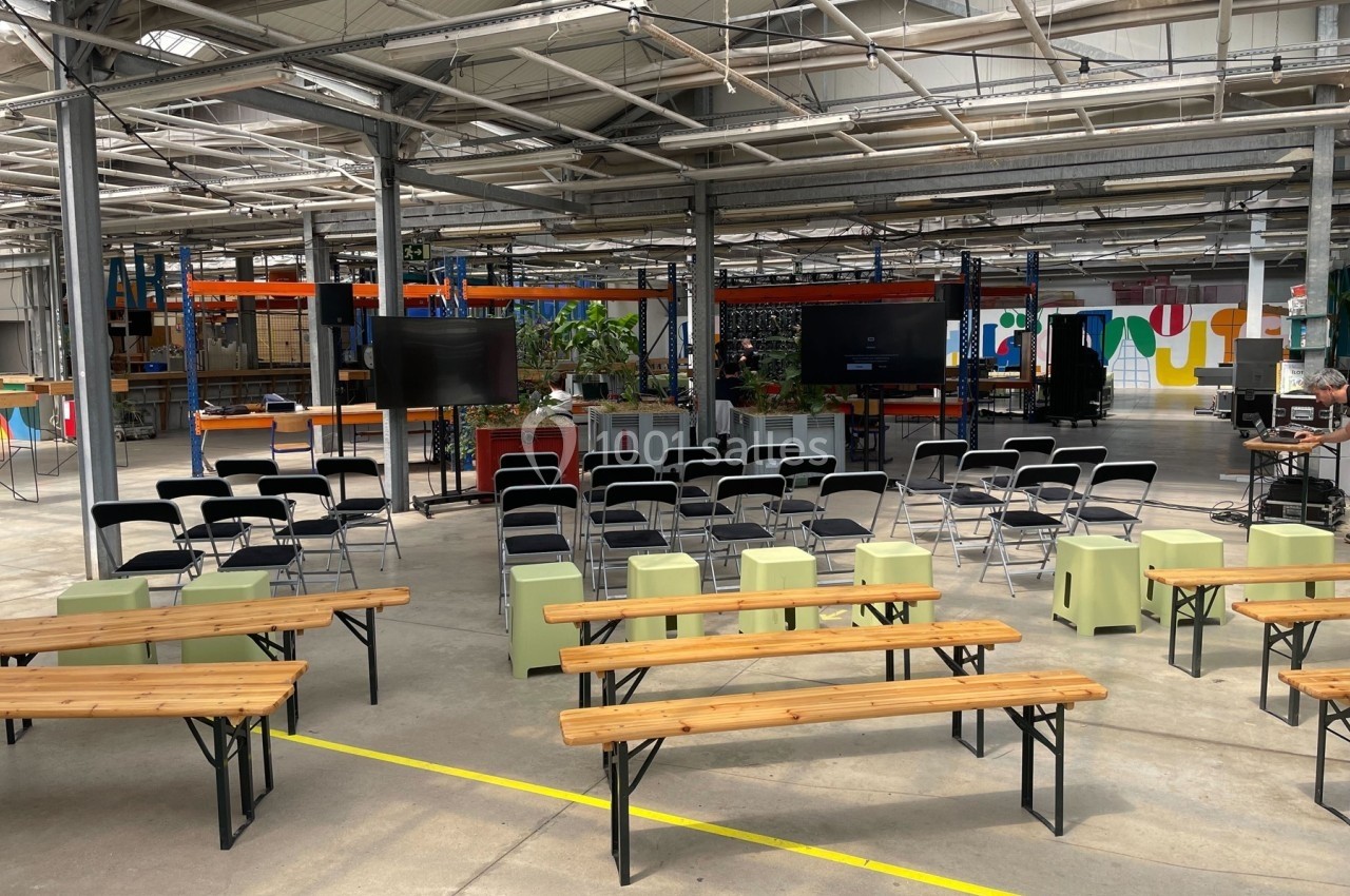 Salle industrielle aménagée avec des bancs, des chaises et des écrans pour un événement ou une présentation.