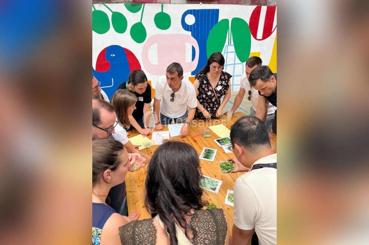 Un groupe de personnes discute autour d'une table en bois, avec des documents et des plantes, devant une fresque colorée.