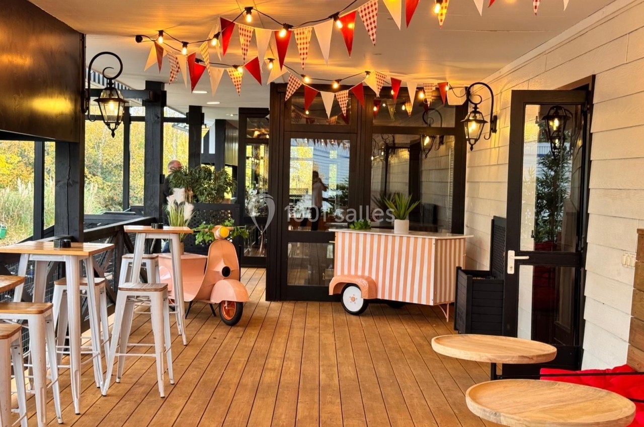 Terrasse en bois décorée de guirlandes lumineuses et fanions, avec tables hautes, scooter vintage et chariot rayé.