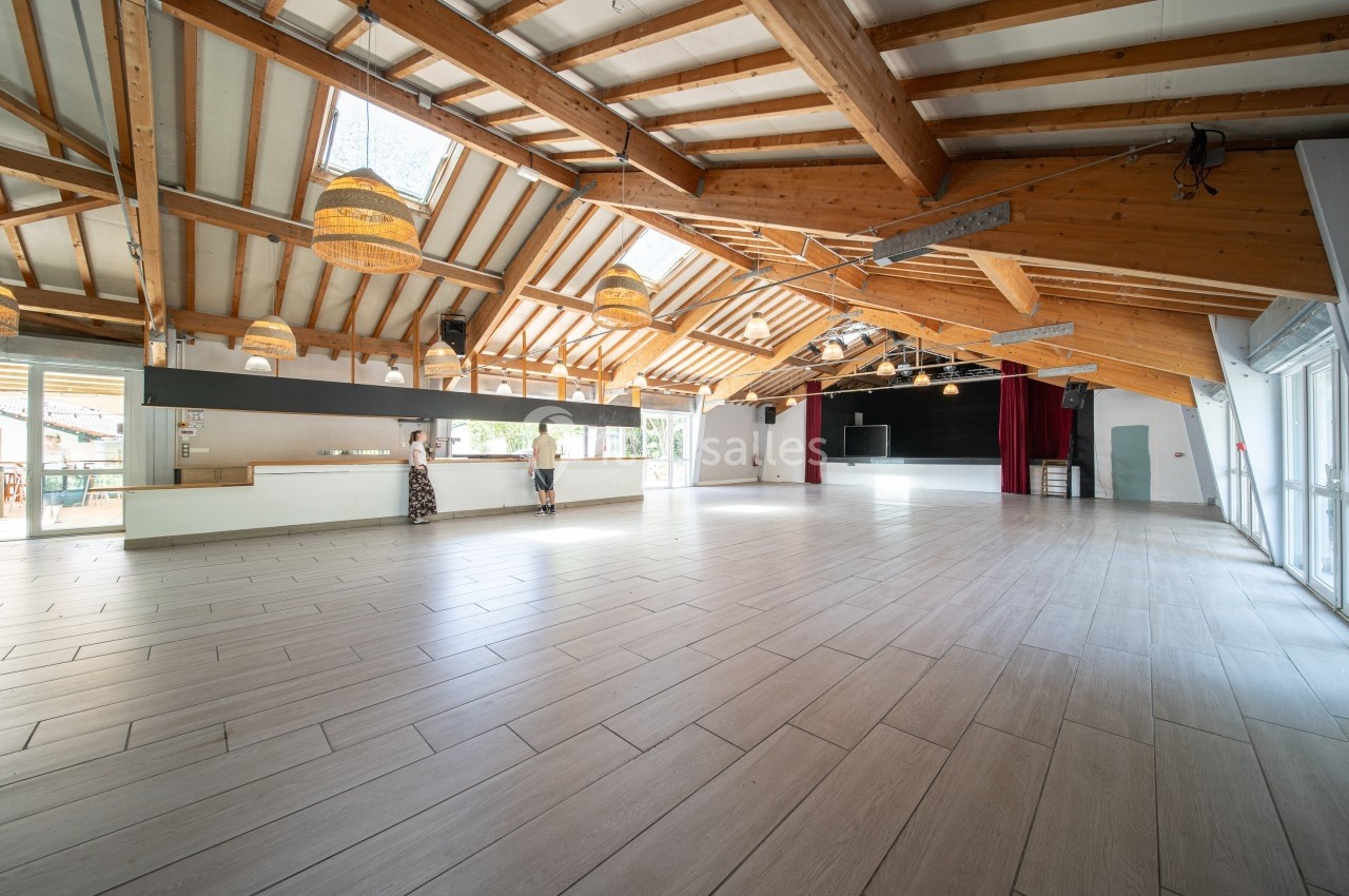 Grande salle lumineuse avec toit en bois, sol carrelé, bar à gauche et scène avec rideaux rouges au fond.
