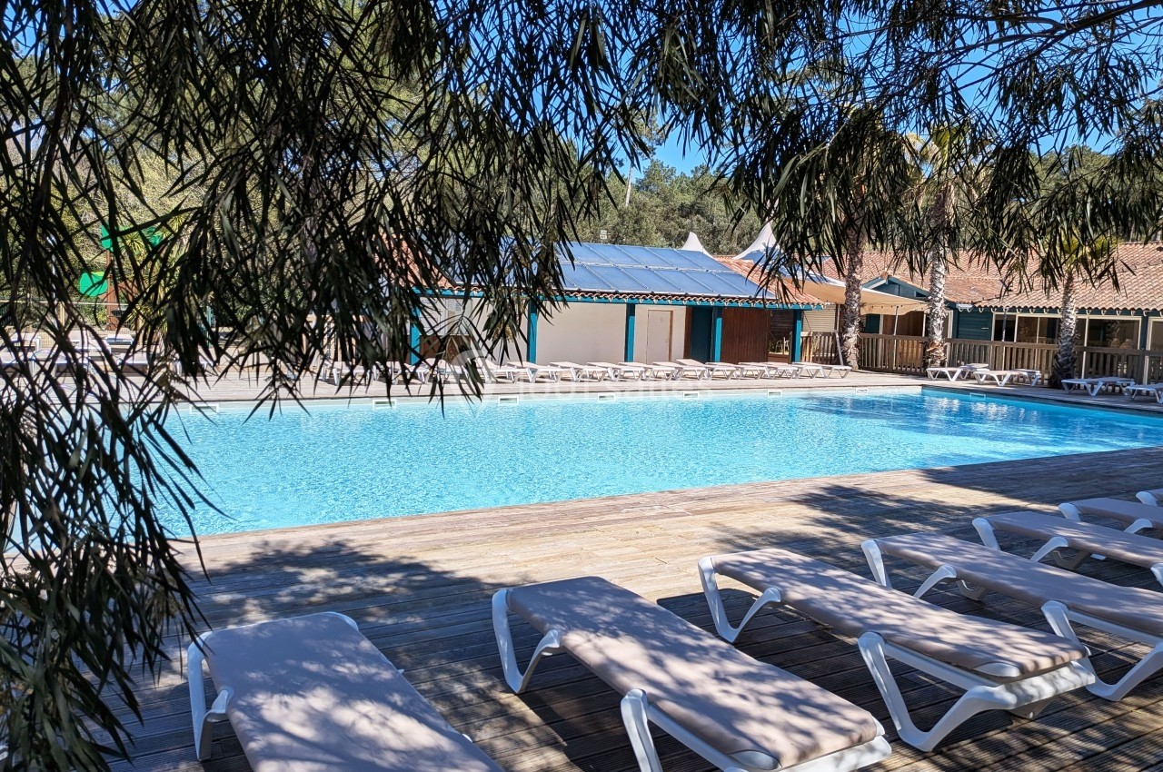 Piscine extérieure entourée de transats sur une terrasse en bois, avec des bâtiments en arrière-plan.
