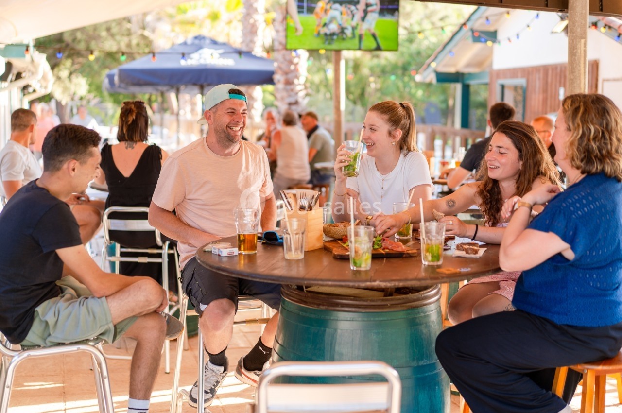 Groupe d'amis souriants partageant un repas et des boissons autour d'une table en terrasse ensoleillée.