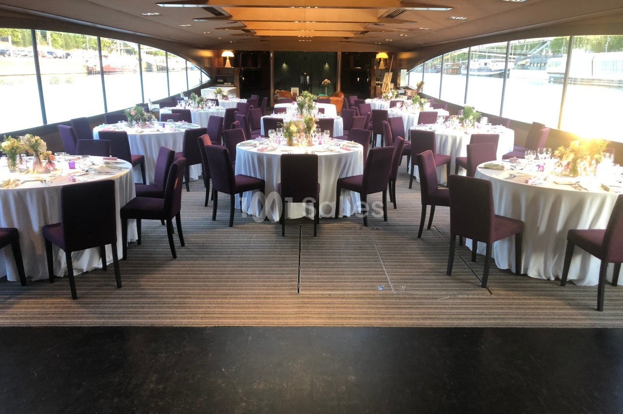 Salle de réception élégante avec tables rondes dressées, nappes blanches et chaises violettes, éclairée par la lumière…