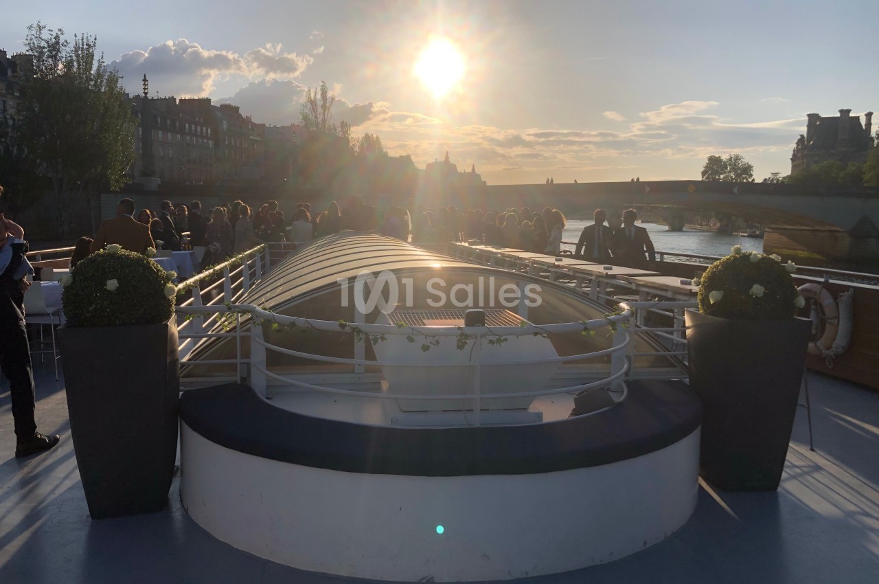 Pont d'un bateau au coucher du soleil, avec des passagers rassemblés et un pont visible à l'arrière-plan.