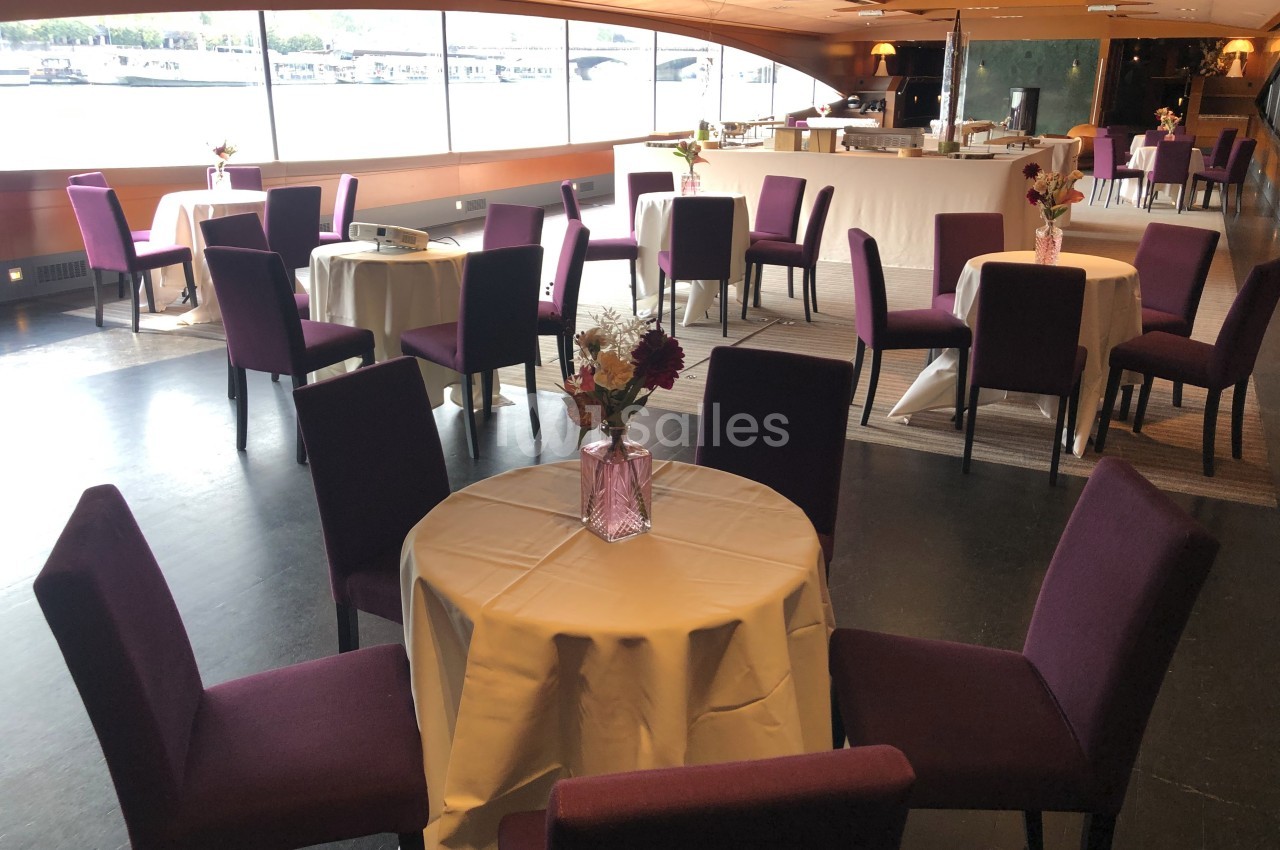Salle aménagée avec des tables rondes nappées et des chaises violettes, offrant une vue sur un paysage fluvial.