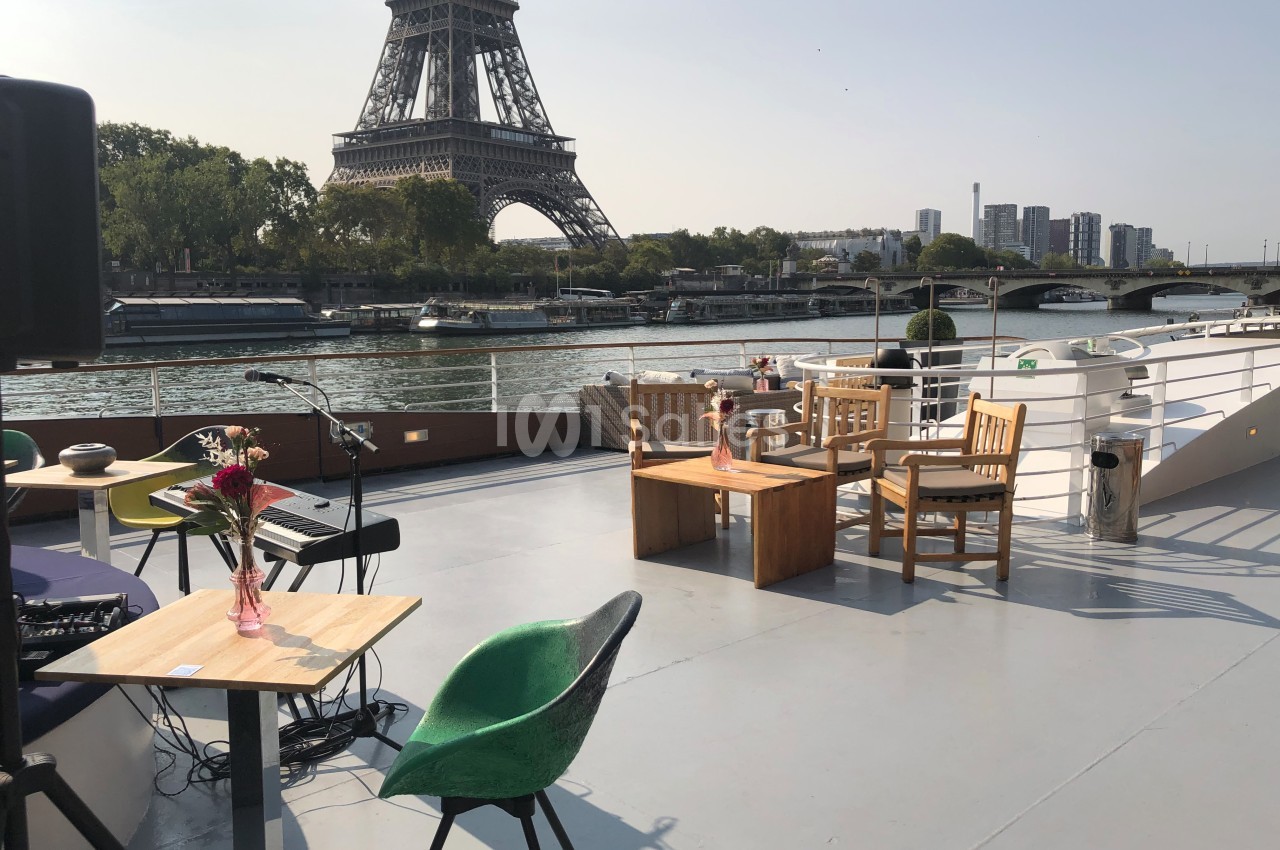 Terrasse d'un bateau avec mobilier en bois et vue sur la Tour Eiffel et la Seine par temps clair.