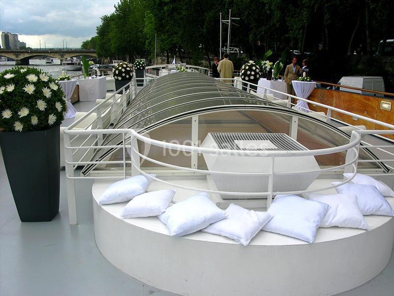Pont supérieur d'un bateau avec des coussins blancs, des décorations florales et une vue sur un quai arboré.