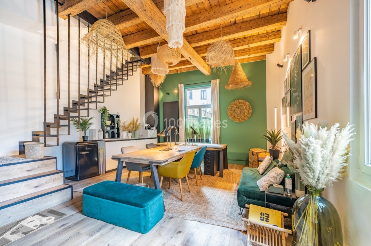 Espace de travail lumineux avec poutres apparentes, mobilier coloré, plantes décoratives et grande fenêtre.