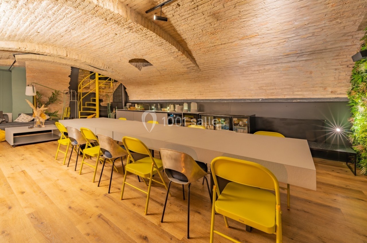 Salle voûtée avec longue table entourée de chaises jaunes, escalier en colimaçon et décor végétal en arrière-plan.