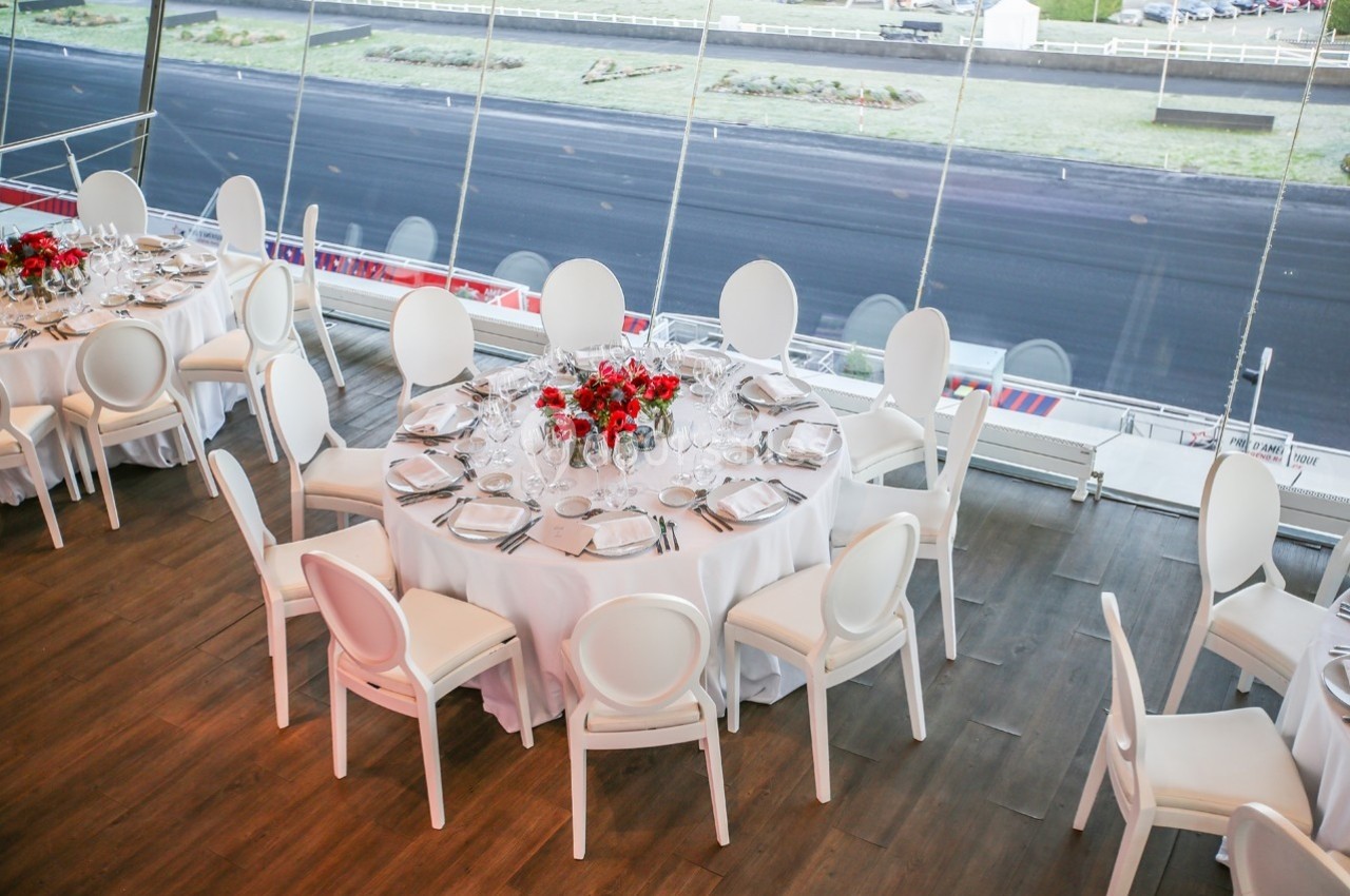 Tables rondes dressées avec nappes blanches, chaises blanches et bouquets rouges, dans une salle avec vue sur une piste…