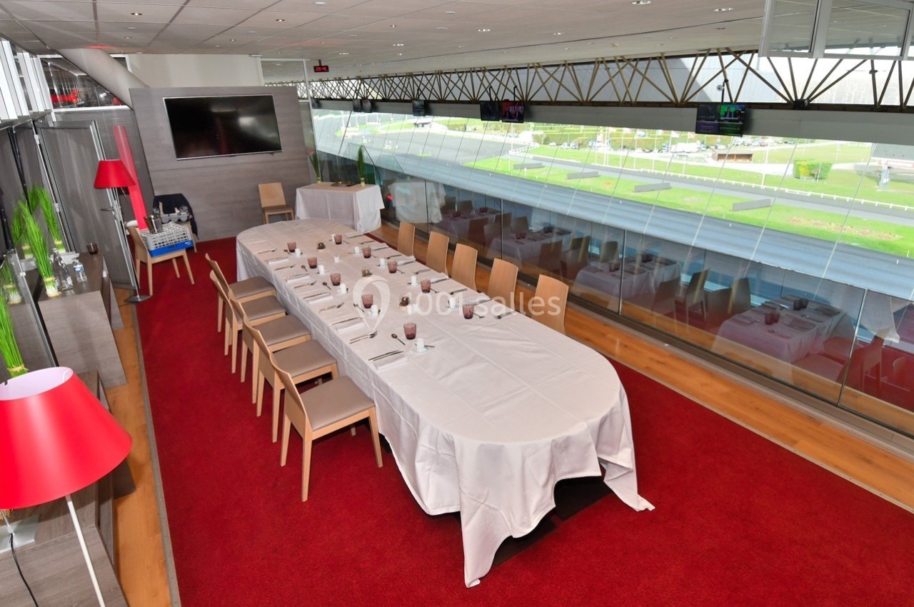Salle de réception avec une grande table dressée, nappée de blanc, entourée de chaises, et vue sur l'extérieur vitré.